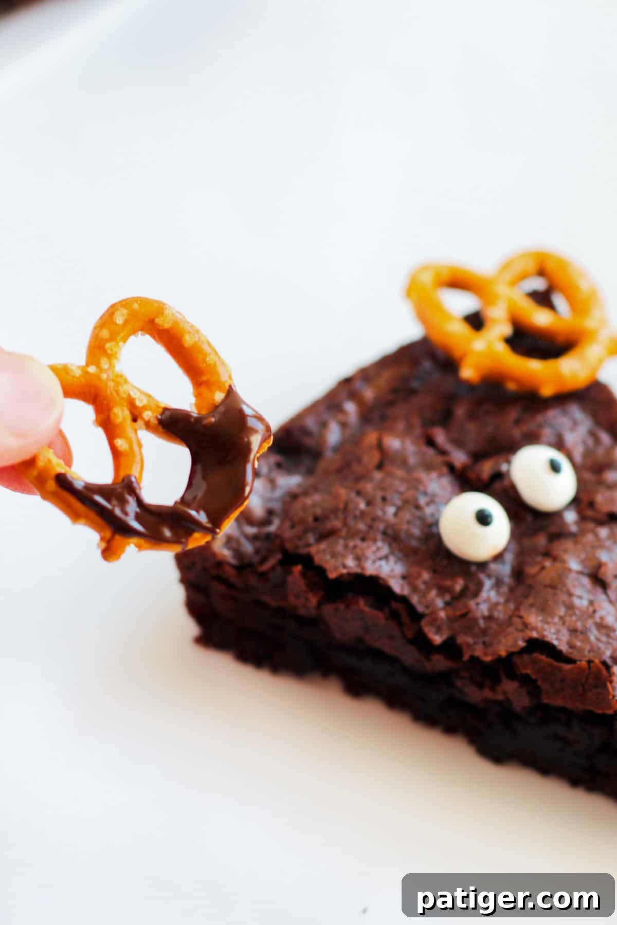 A close-up view of a hand carefully placing a pretzel twist, coated with melted chocolate, onto a brownie above two candy eyes, demonstrating the step-by-step decoration process.
