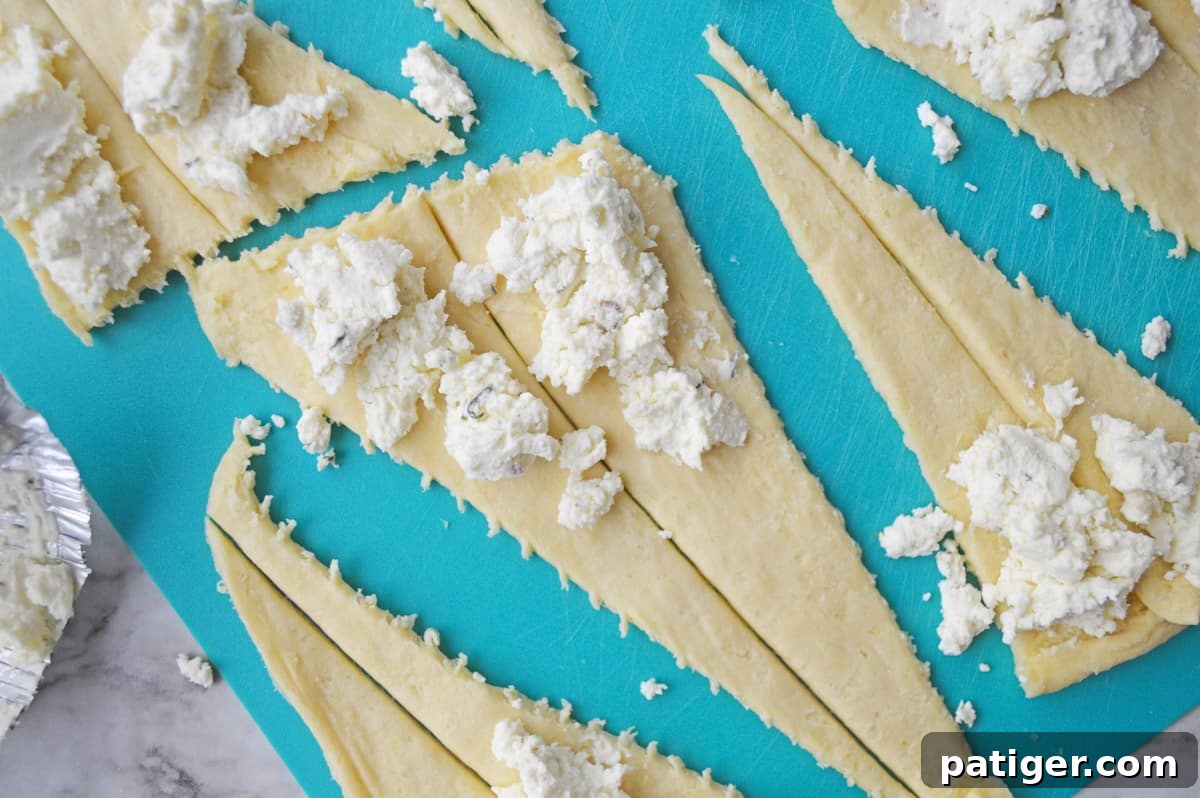 Close-up shot of Boursin cheese being carefully spread onto the wide end of each crescent dough triangle, preparing it for rolling.