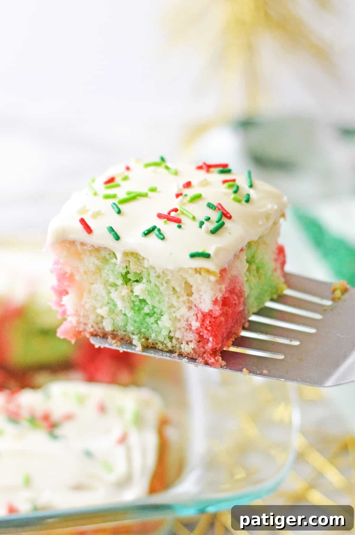 Square slice of red and green jello poke cake being lifted out of baking dish with metal spatula.