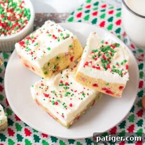 Thick Christmas sugar cookie bars with sprinkles in the dough and topped with vanilla frosting and more sprinkles. A glass of milk and bowl of holiday chips can be seen around them.