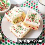 Thick Christmas sugar cookie bars with sprinkles in the dough and topped with vanilla frosting and more sprinkles. A glass of milk and bowl of holiday chips can be seen around them.