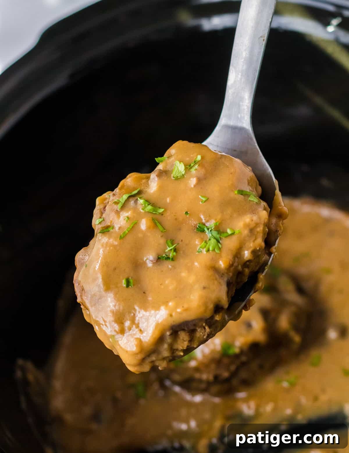 Tender Slow Cooker Salisbury Steaks 8 Serving spoon lifting gravy smothered salisbury steak patty out of crockpot.