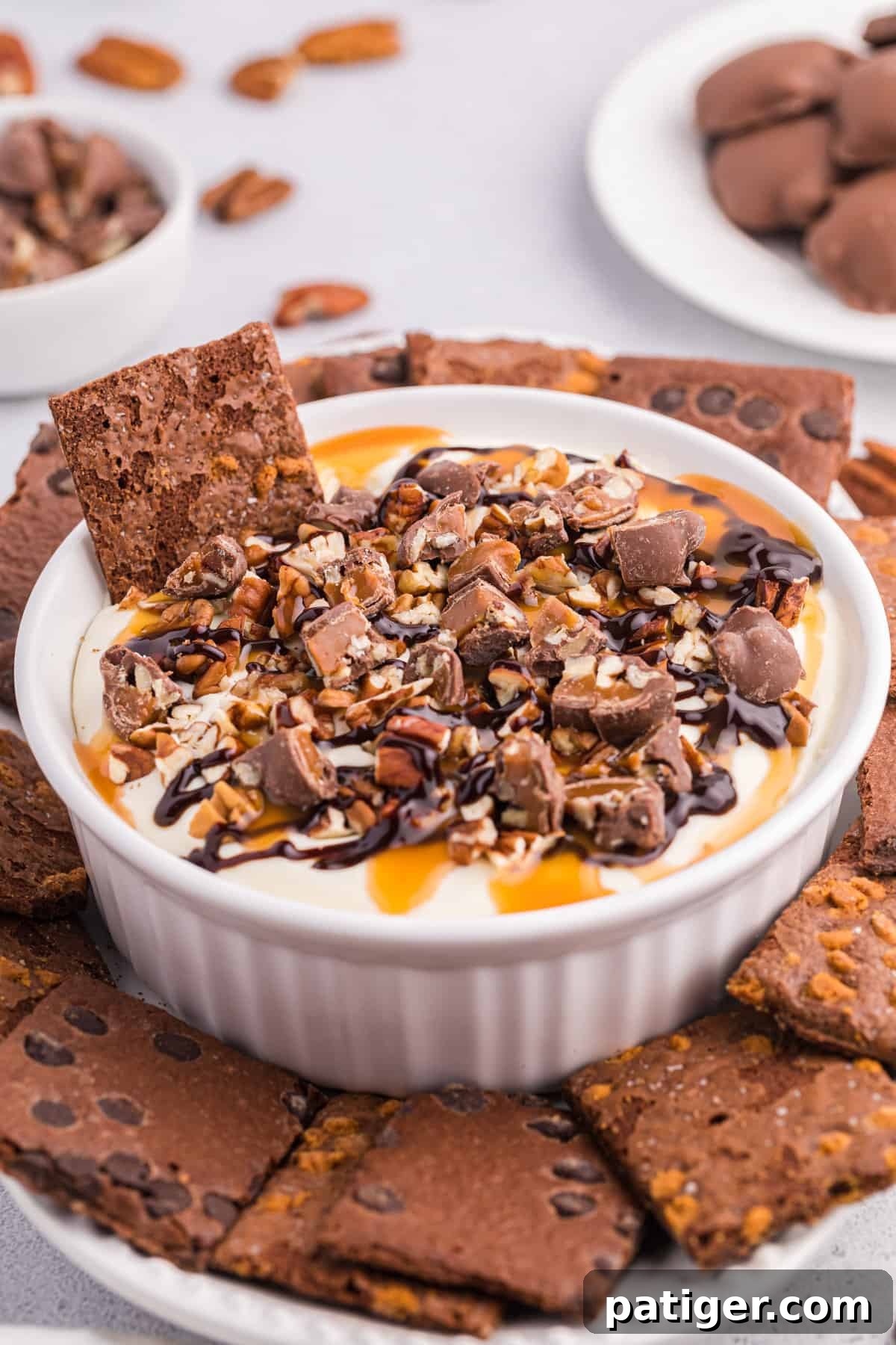 No-bake turtle cheesecake dip in a white serving bowl topped with caramel, pecans, chopped turtle candies, and served with brownie brittle for dipping.