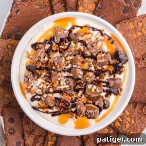 No-bake turtle cheesecake dip in a white serving bowl topped with caramel, pecans, chopped turtle candies, and served with brownie brittle for dipping.