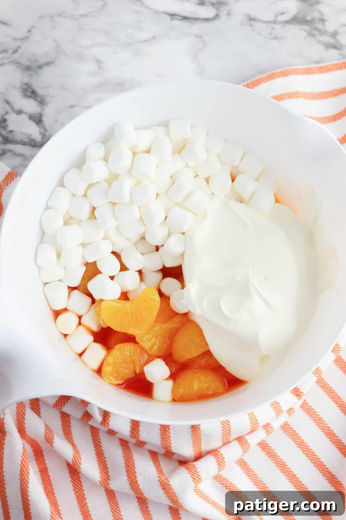 Mandarin oranges, mini marshmallows, and whipped topping being gently folded into the partially set orange jello in a mixing bowl.
