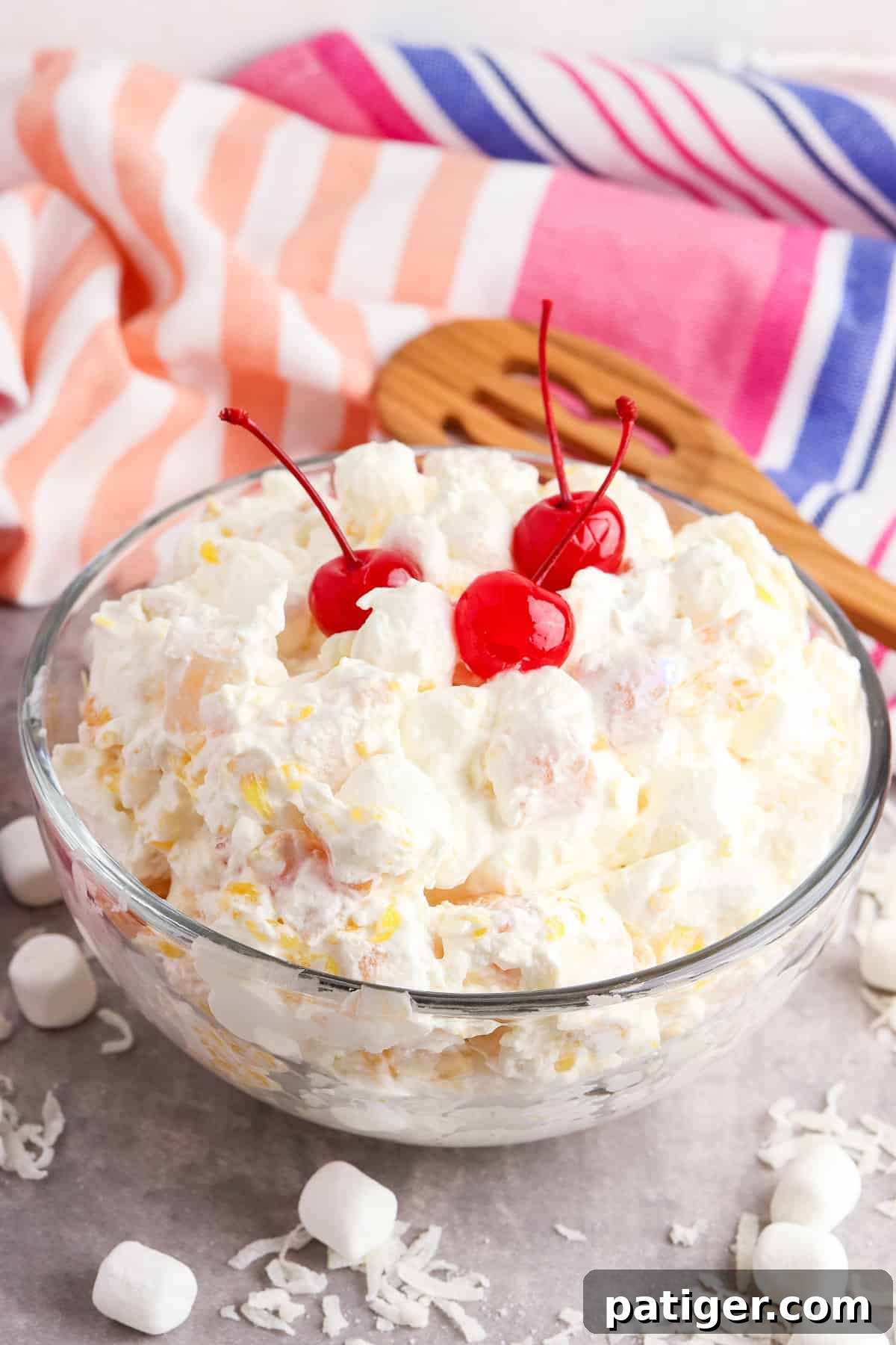 Cloud Nine Fruit Salad 2 A large glass bowl filled with colorful marshmallow fruit salad, garnished with a wooden slotted spoon and set against a patterned linen.