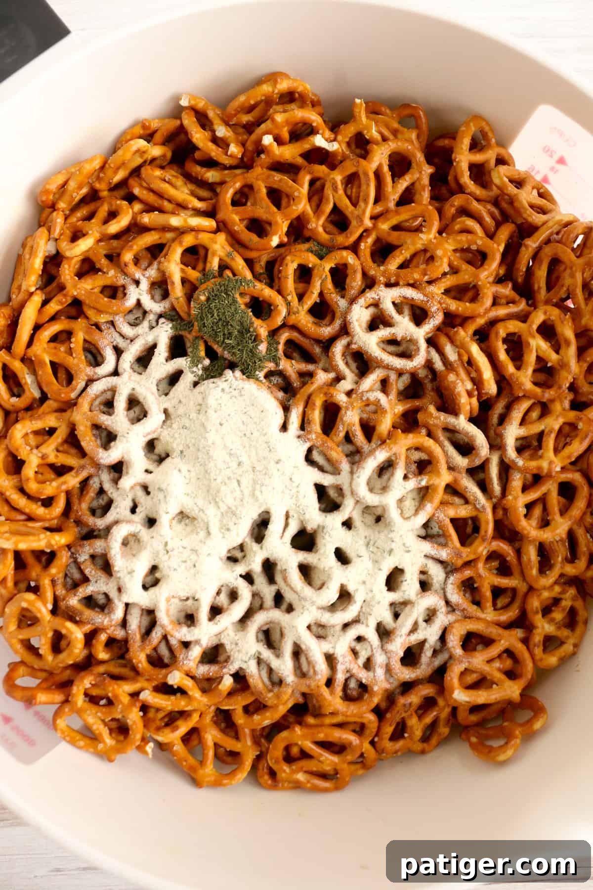 Dry ranch mix and other seasonings are generously sprinkled over pretzel twists in a large mixing bowl, ready for shaking.