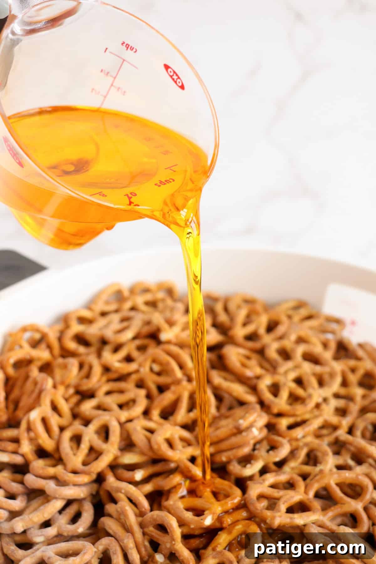 Golden popcorn oil is being poured into a bowl of seasoned pretzels, preparing for the next mixing stage.