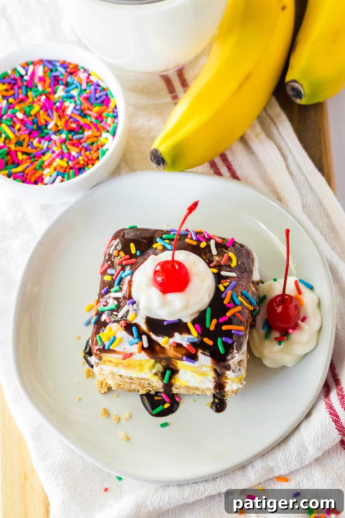 Square piece of no bake banana split cake on white plate with whipped cream and a cherry. Rainbow sprinkles and fresh bananas are next to the plate.