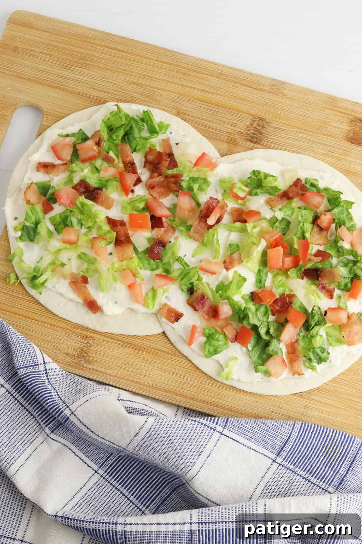 Two flour tortillas layed slightly overlapping on a cutting board. The tortillas are topped with creamy dressing, shredded lettuce, diced tomatoes, and chopped bacon, ready to be rolled into delicious BLT pinwheels.
