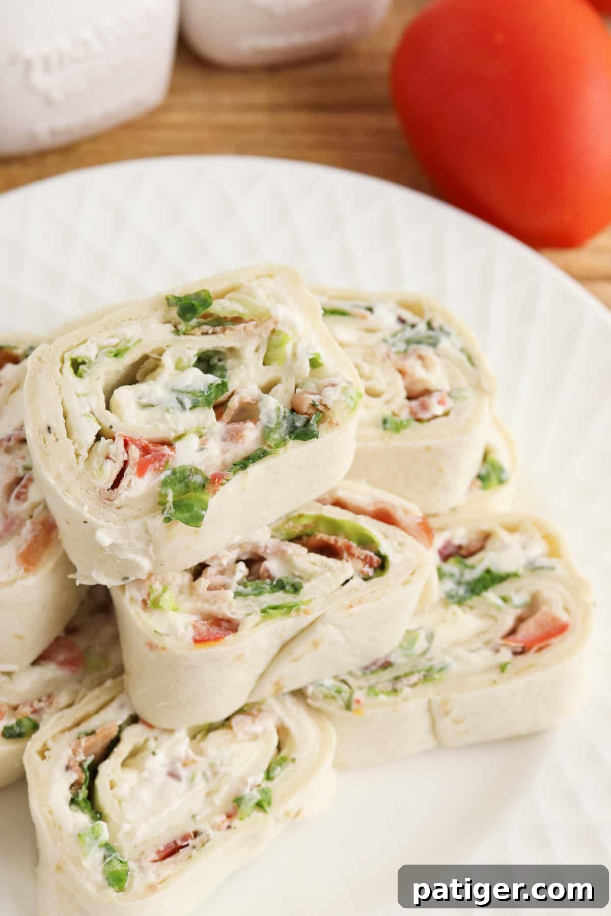 Ranch BLT Pinwheels made with flour tortillas, bacon, lettuce, tomatoes, and ranch dressing stacked on a white plate with a plum tomato next to it. These vibrant pinwheels are a perfect, colorful appetizer.
