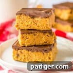 Three no-bake chocolate peanut butter bars stacked on a plate with milk and more bars behind them.