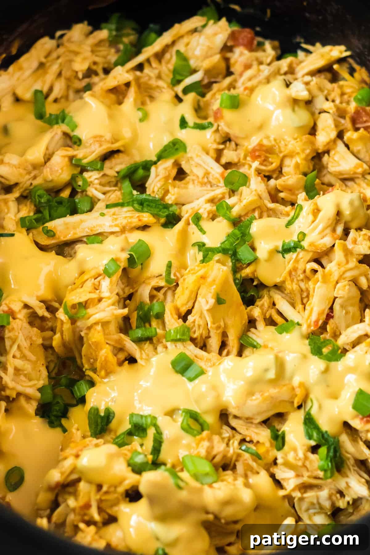 Close-up of cheesy shredded chicken simmering in a crockpot, generously topped with melted queso and fresh chopped green onions.