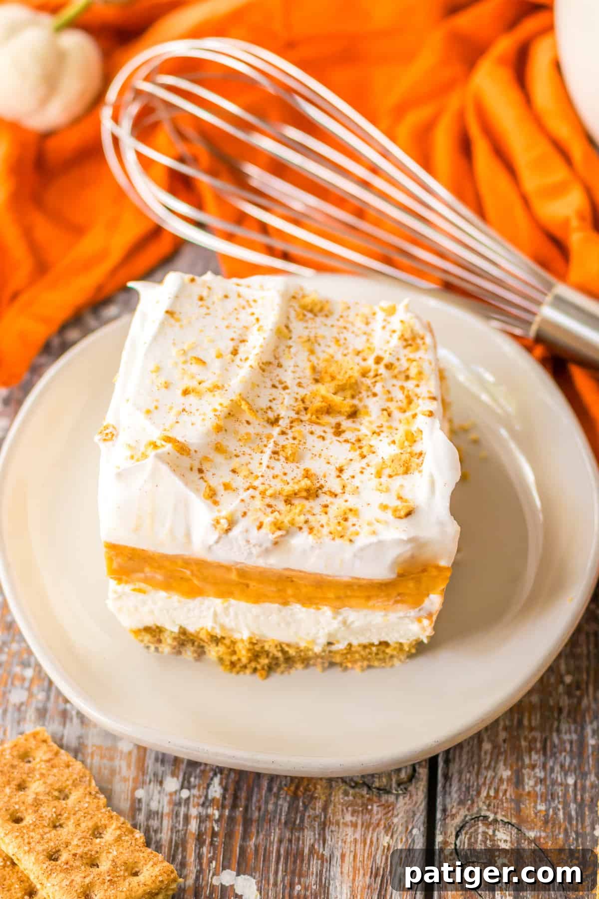 A square piece of layered pumpkin dessert on a white plate, showcasing the distinct layers, accompanied by a wire whisk and orange linen.