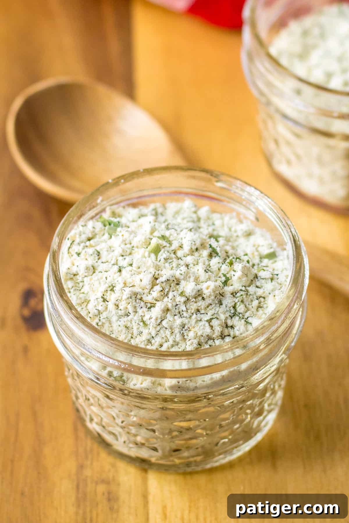 Delicious Homemade Ranch Seasoning 2 Homemade ranch seasoning mix in a glass jar with a wooden spoon next to it, against a rustic backdrop.