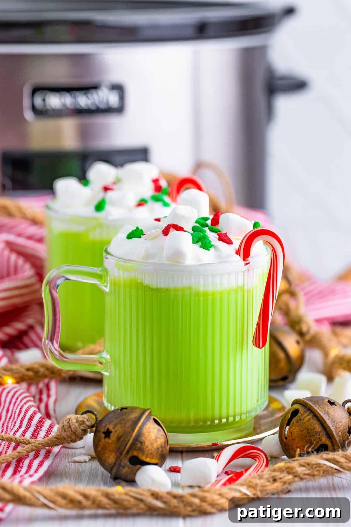 Slow cooker grinch hot chocolate in two glass mugs with whipped cream, mini marshmallows, christmas sprinkles, and mini candy canes. A crockpot is in the background.
