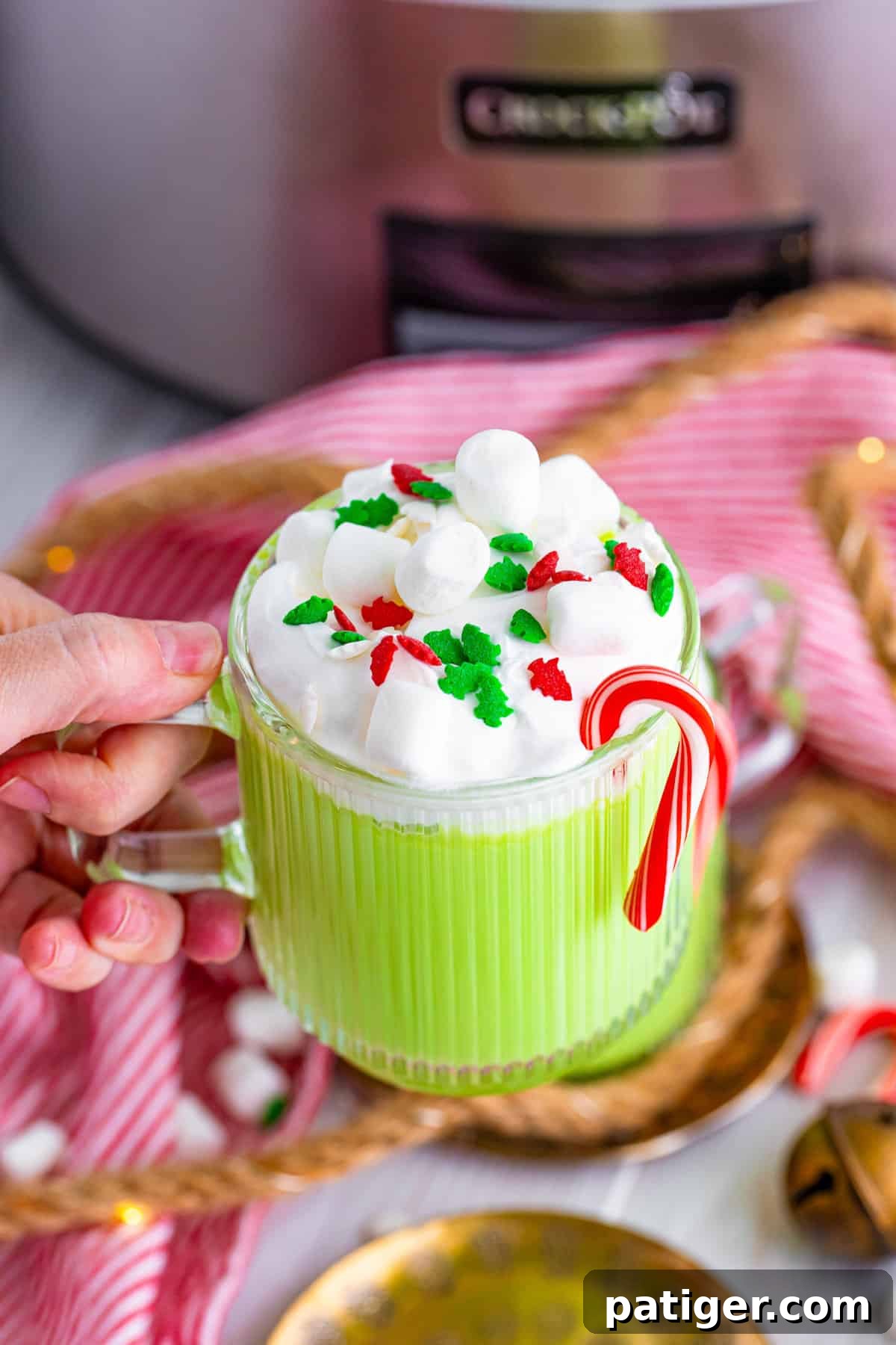 Hand holidaying mug of Grinch hot chocolate with a slow cooker in the background.
