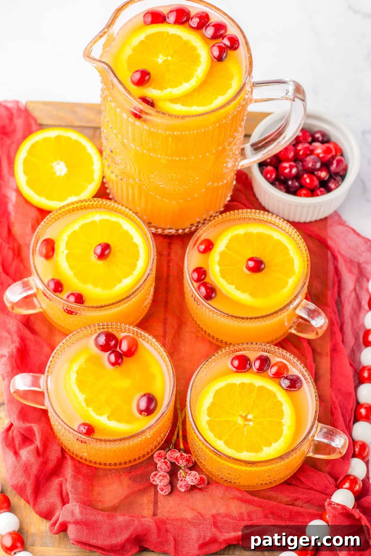A glass pitcher filled with non-alcoholic Christmas punch, beautifully adorned with fresh orange slices and cranberries, alongside four individual glasses.