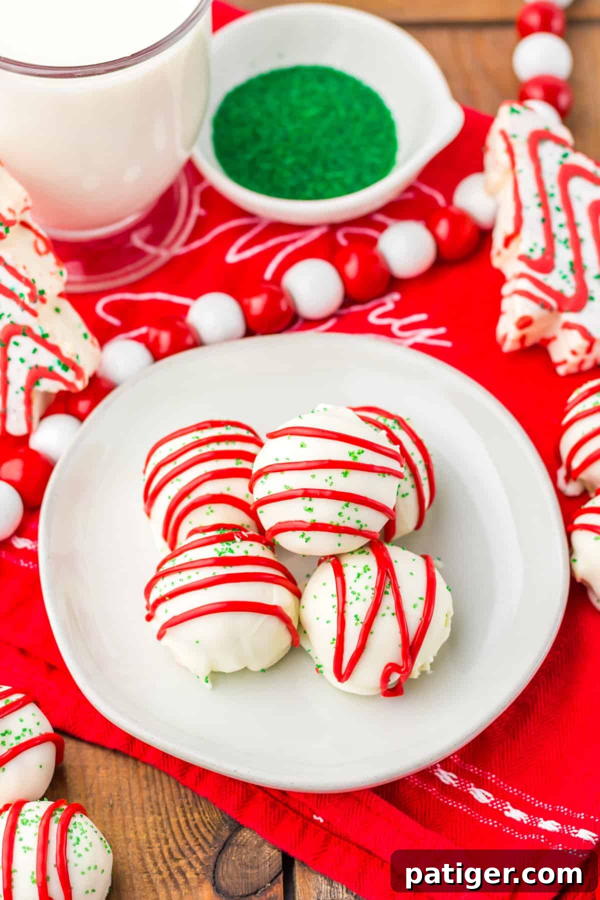 Little Debbie Christmas Tree Cake Balls arranged on a white plate, surrounded by green sugar sprinkles, whole Little Debbie Christmas Tree Snack Cakes, and a glass of milk.