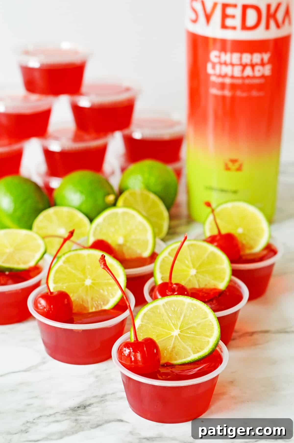 Cherry Limeade Jello Shots made with red jello and garnished with lime and maraschino cherries. Fresh limes, shots covered with lids, and a bottle of SVEDKA cherry limeade vodka are in the background.