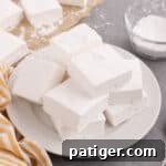 Fluffy homemade marshmallows piled on white plate coated in powdered sugar.