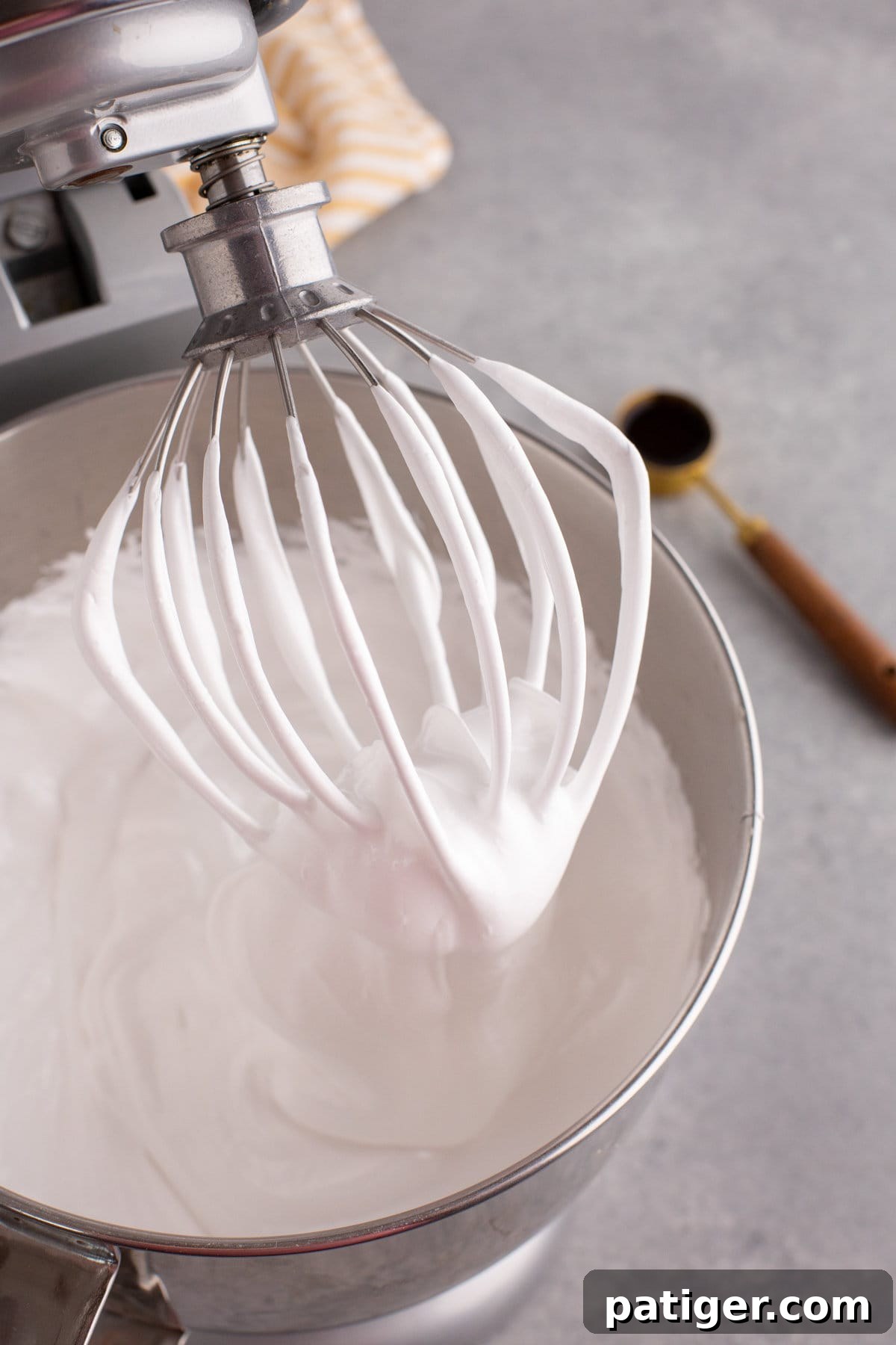 Stand mixer head tilted back to show whisk attachment with white fluffy marshmallow mixture on it.