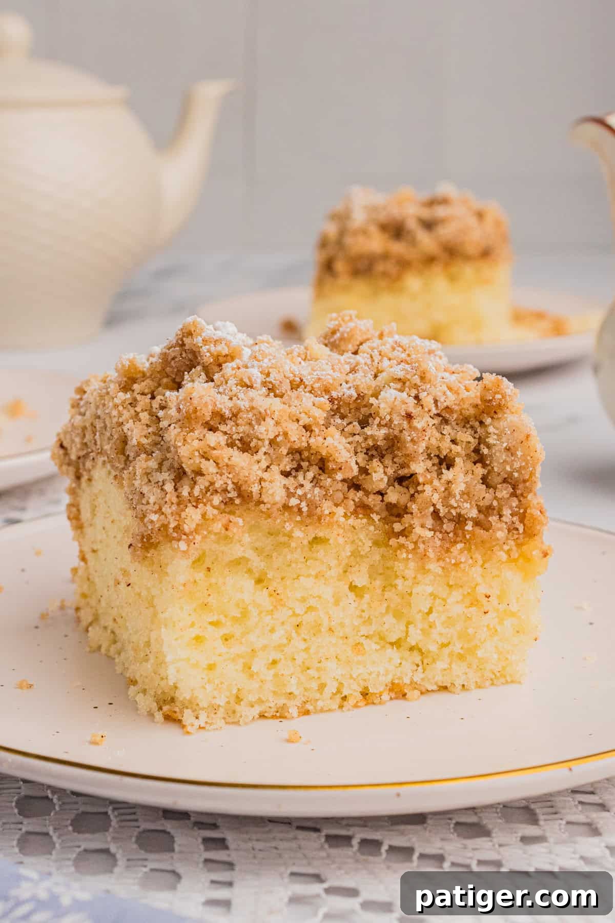A perfectly sliced piece of New York style crumb cake featuring a thick, golden crumb topping. Another slice and a vintage teapot are blurred in the background, suggesting a cozy setting.