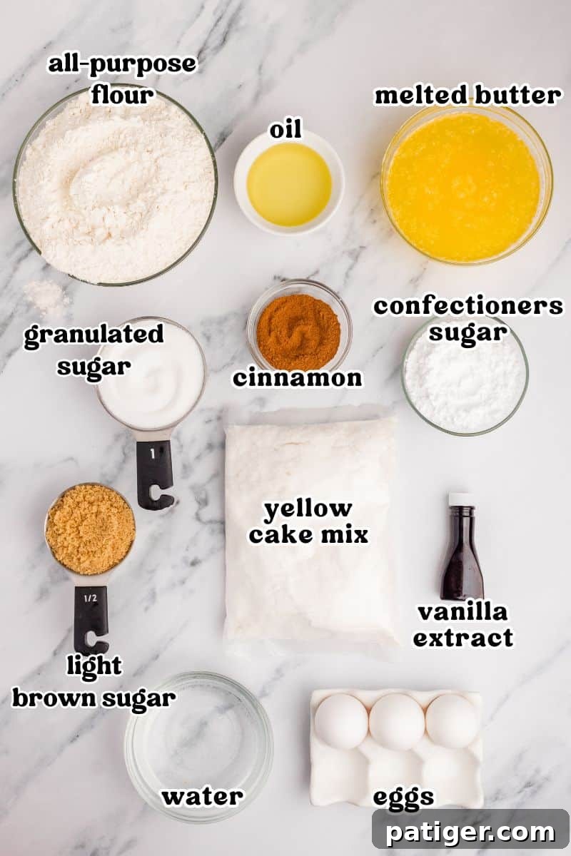 A collection of ingredients neatly arranged on a countertop, including a box of yellow cake mix, bottles of oil and vanilla extract, a stick of butter, granulated and light brown sugars, confectioners sugar, eggs, water, and a jar of ground cinnamon.