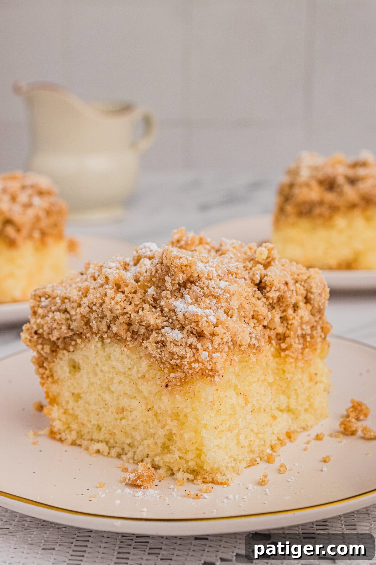 A close-up of a slice of New York style crumb cake, with its distinct layers of moist cake and thick crumb topping visible. A teapot is visible in the background, out of focus.