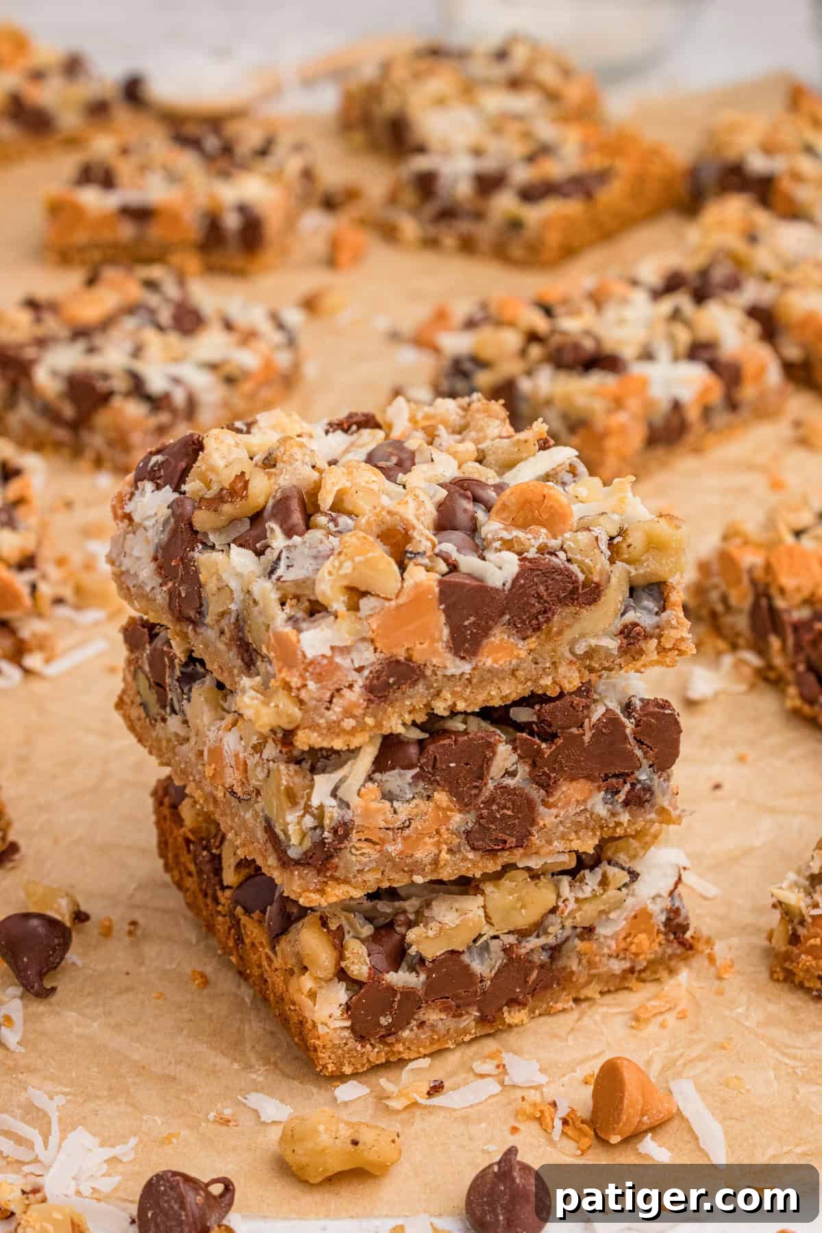 7 Layer bars with chocolate chips, butterscotch chips, shredded coconut, and chopped walnuts over a graham cracker crust. Three of the cookie bars are stacked on top of each other with additional bars in the background.