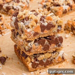 7 Layer bars with chocolate chips, butterscotch chips, shredded coconut, and chopped walnuts over a graham cracker crust. Three of the cookie bars are stacked on top of each other with additional bars in the background.