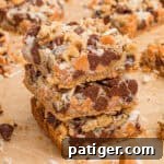 7 Layer bars with chocolate chips, butterscotch chips, shredded coconut, and chopped walnuts over a graham cracker crust. Three of the cookie bars are stacked on top of each other with additional bars in the background.