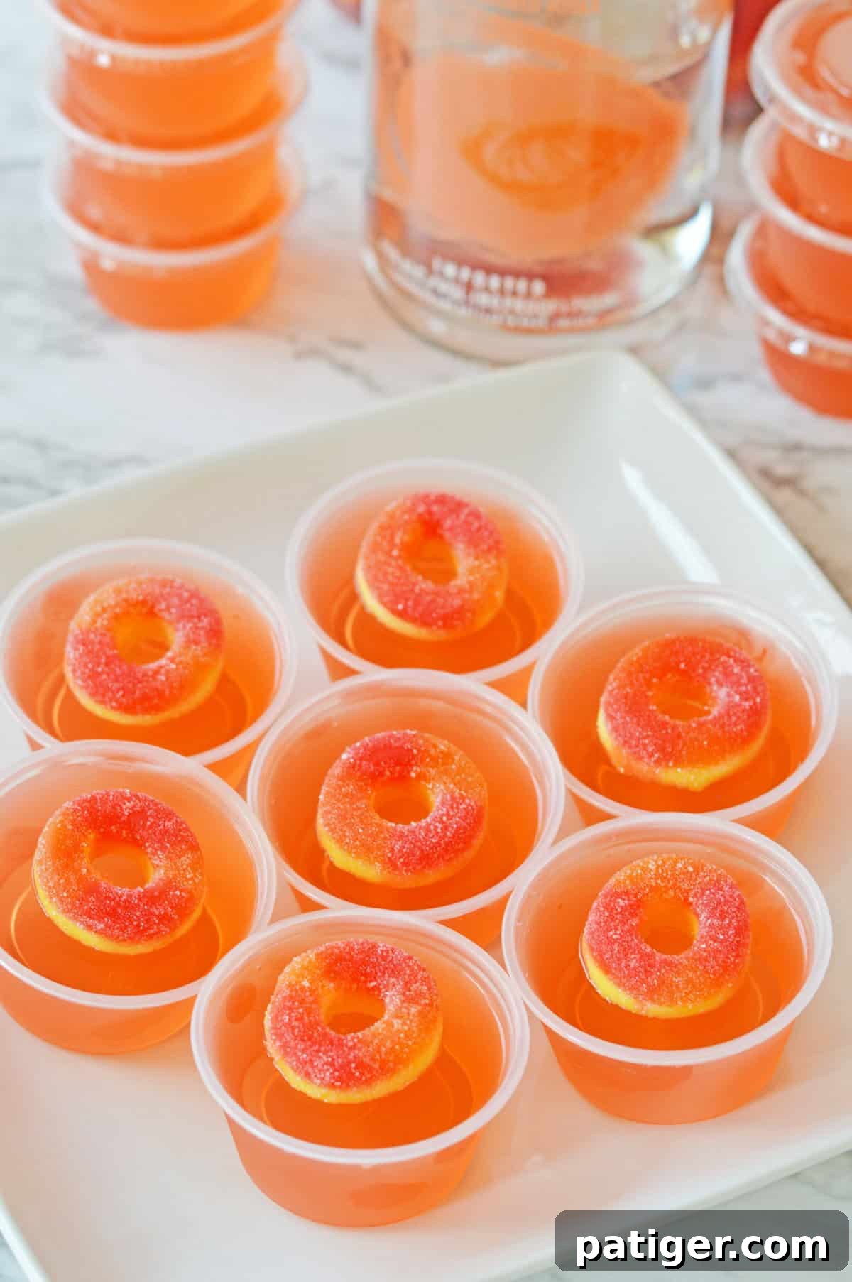 Peach jello shots with peach vodka in plastic condiment cups on a white plate with peach vodka and more jello shots in the background.