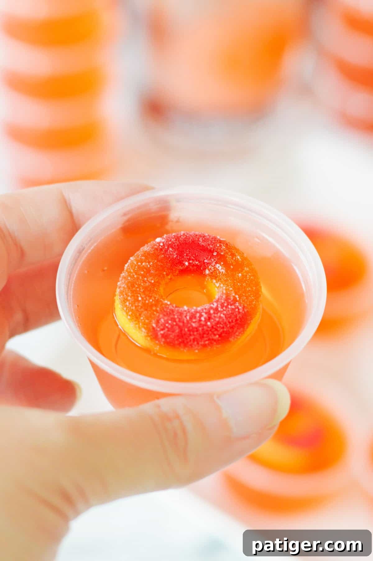 Peach vodka jello shot topped with peach candy ring in a small plastic cup being held by a thumb and forefinger.