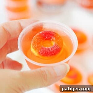 Peach vodka jello shot topped with peach candy ring in a small plastic cup being held by a thumb and forefinger.