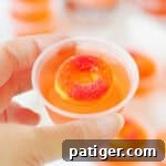 Peach vodka jello shot topped with peach candy ring in a small plastic cup being held by a thumb and forefinger.