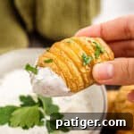 Mini hasselback potato garnished with fresh parsley and dipped in creamy dill dip, on a white plate.