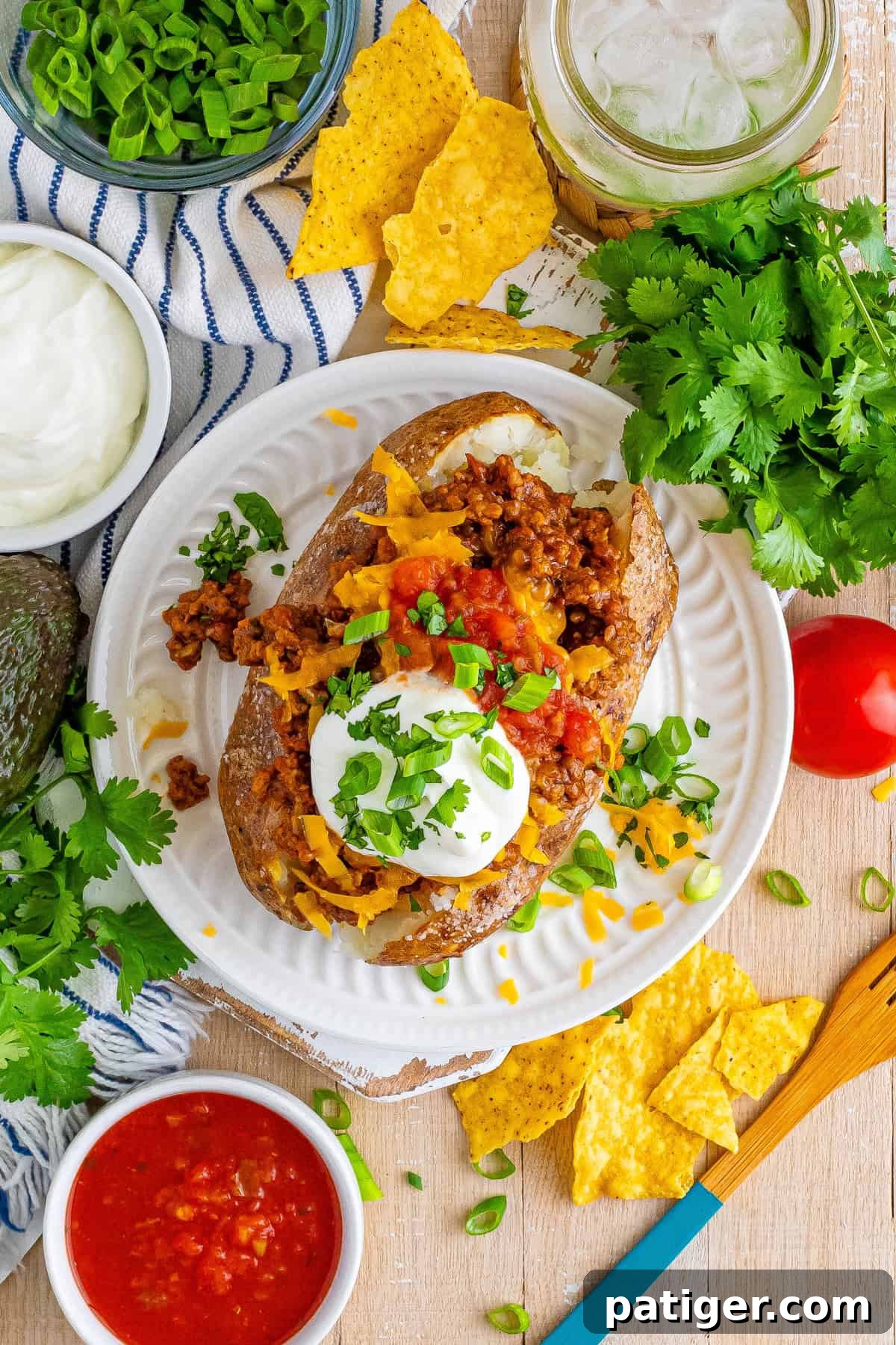 Taco Spud Boats 8 A top-down view of a taco loaded baked potato covered in ground beef, cheese, salsa, sour cream, and green onions, surrounded by taco toppings and tortilla chips on the table.
