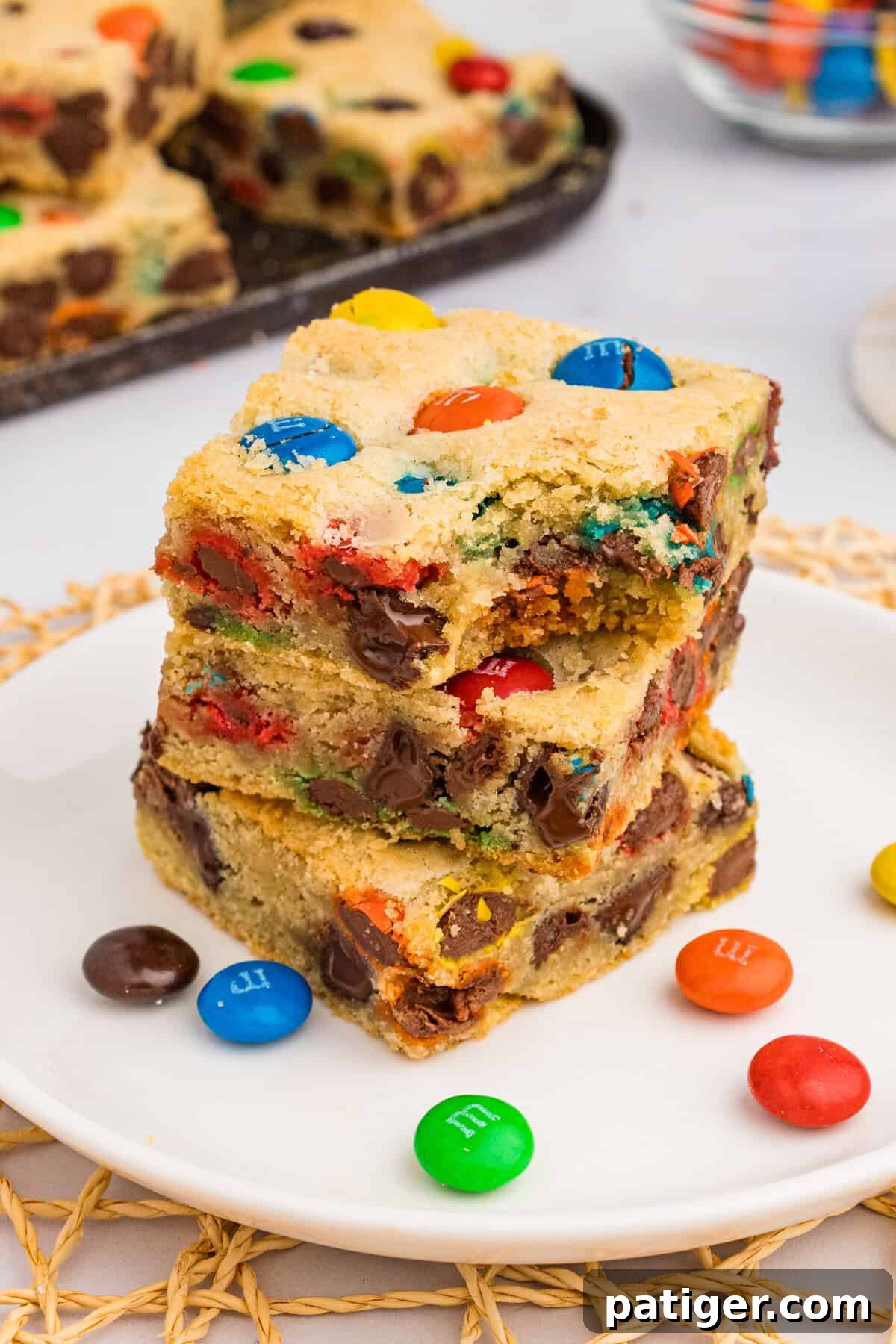 Three thick M&M cookie bars stacked on top of each other, one with a bite taken out, highlighting their thickness and generous M&M distribution and appealing texture.