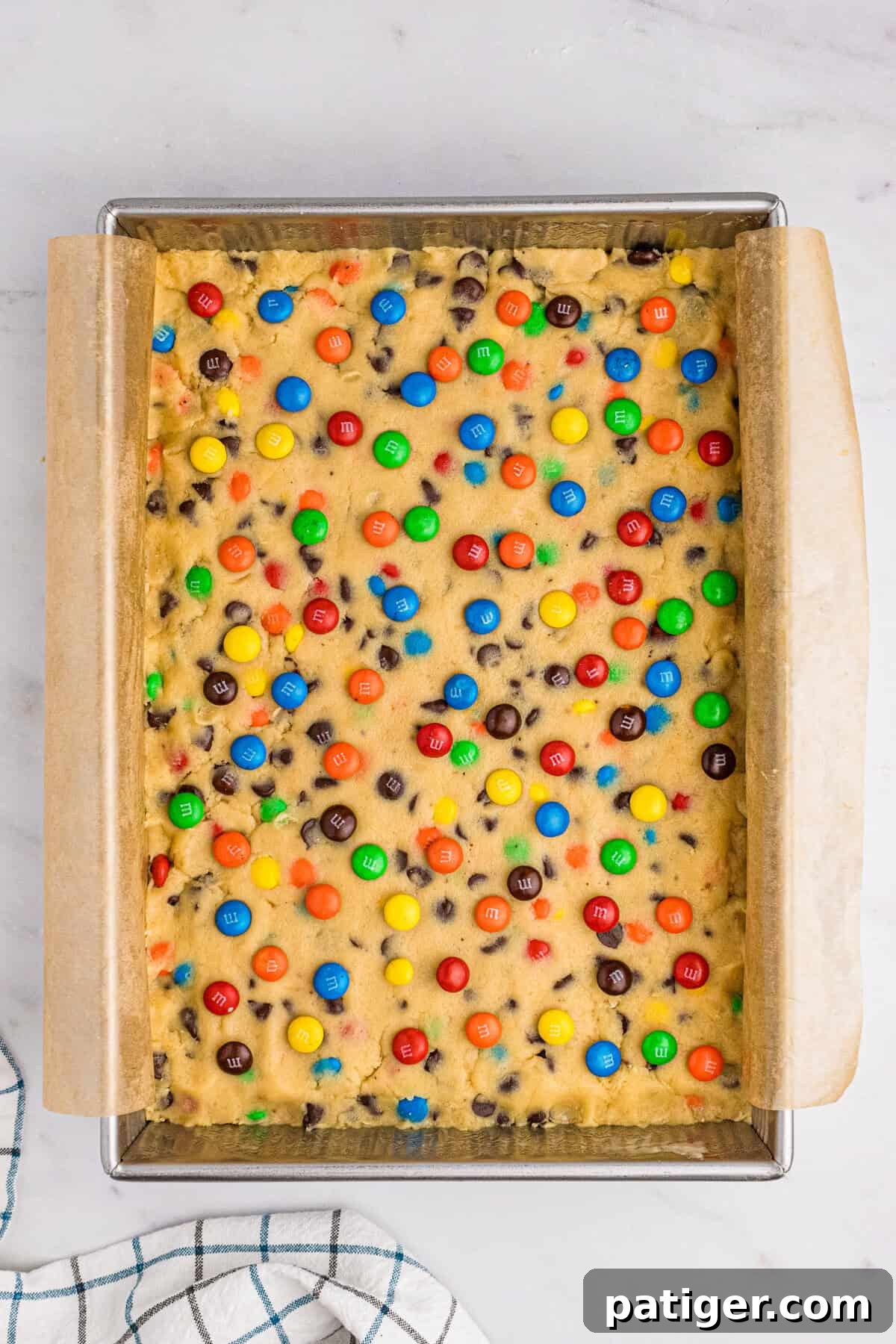 Chocolate chip M&M cookie dough pressed into a parchment-lined 9x13 inch baking pan, generously topped with additional M&Ms before baking.