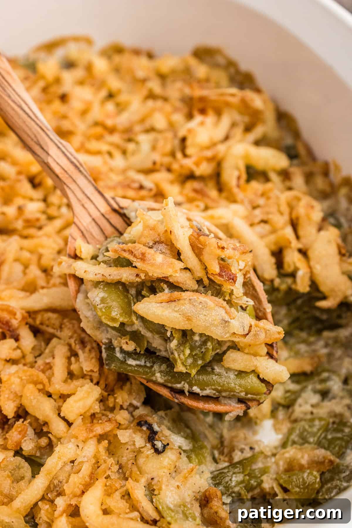 Creamy Green Bean Crunch 2 Spoonful of green bean casserole with Worcestershire sauce and crispy fried onions being served from a casserole dish with a wooden spoon.