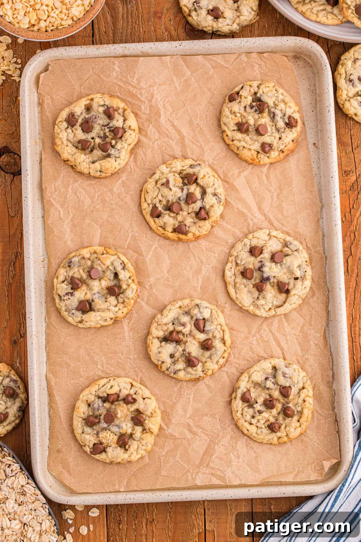 Flat-lay view of Ranger Cookies on a parchment-lined baking sheet, displaying even spacing and a mix of chocolate chip-studded cookies, some with visible cereal flakes, cooling after baking.