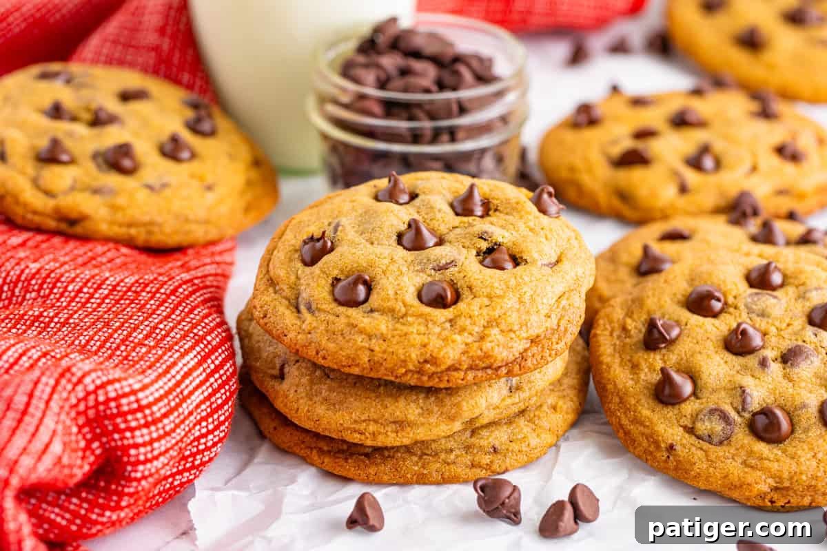 A cozy setup of Mrs. Fields-style cookies next to a glass of milk and a jar of chocolate chips. These cookies are soft, chewy, and a perfect recreation of the Mrs. Fields cookie recipe.