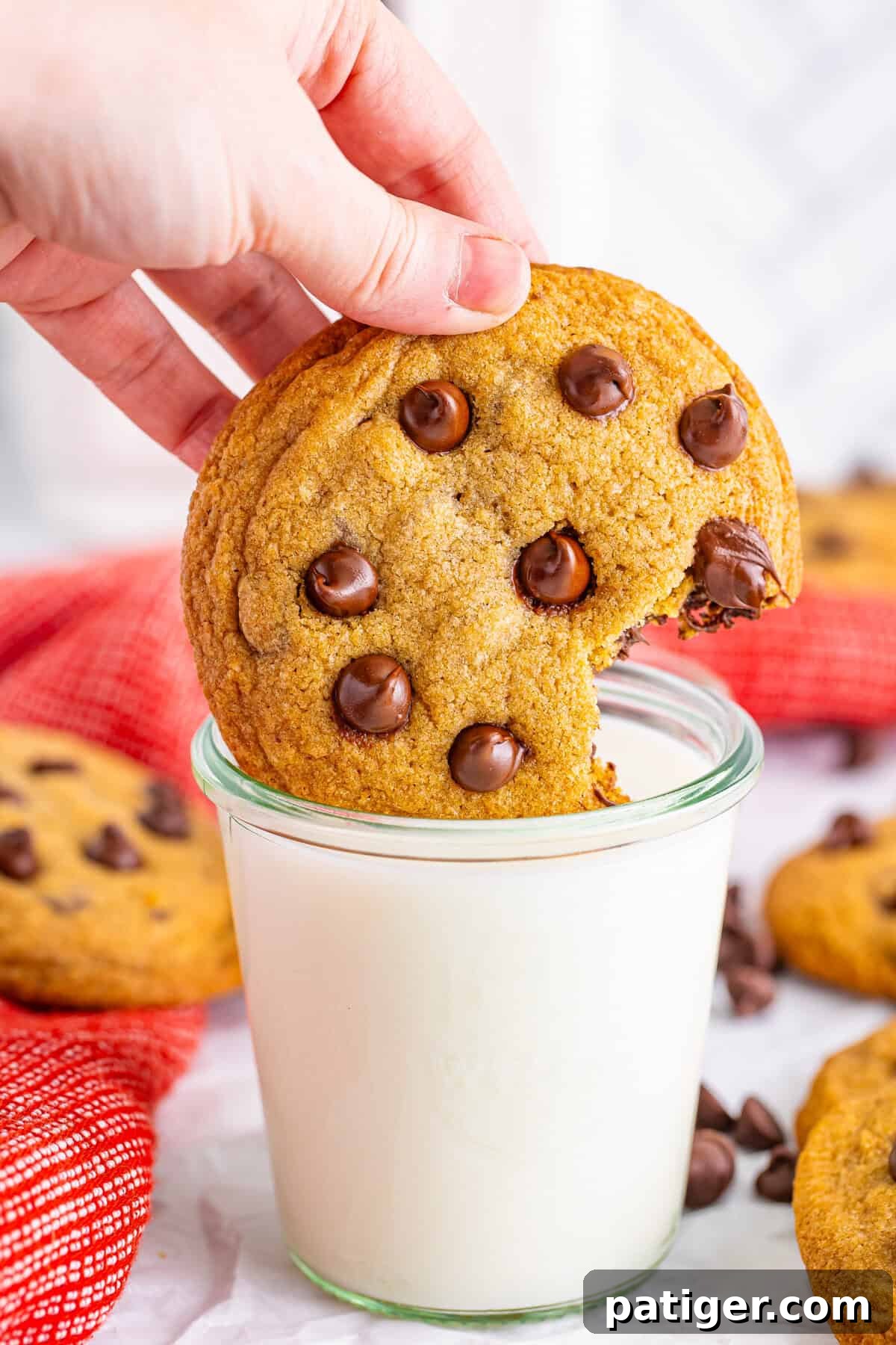 A Mrs. Fields-style chocolate chip cookie being dipped into a glass of milk. The soft, golden-brown cookie is studded with melty chocolate chips, ideal for cookie lovers.