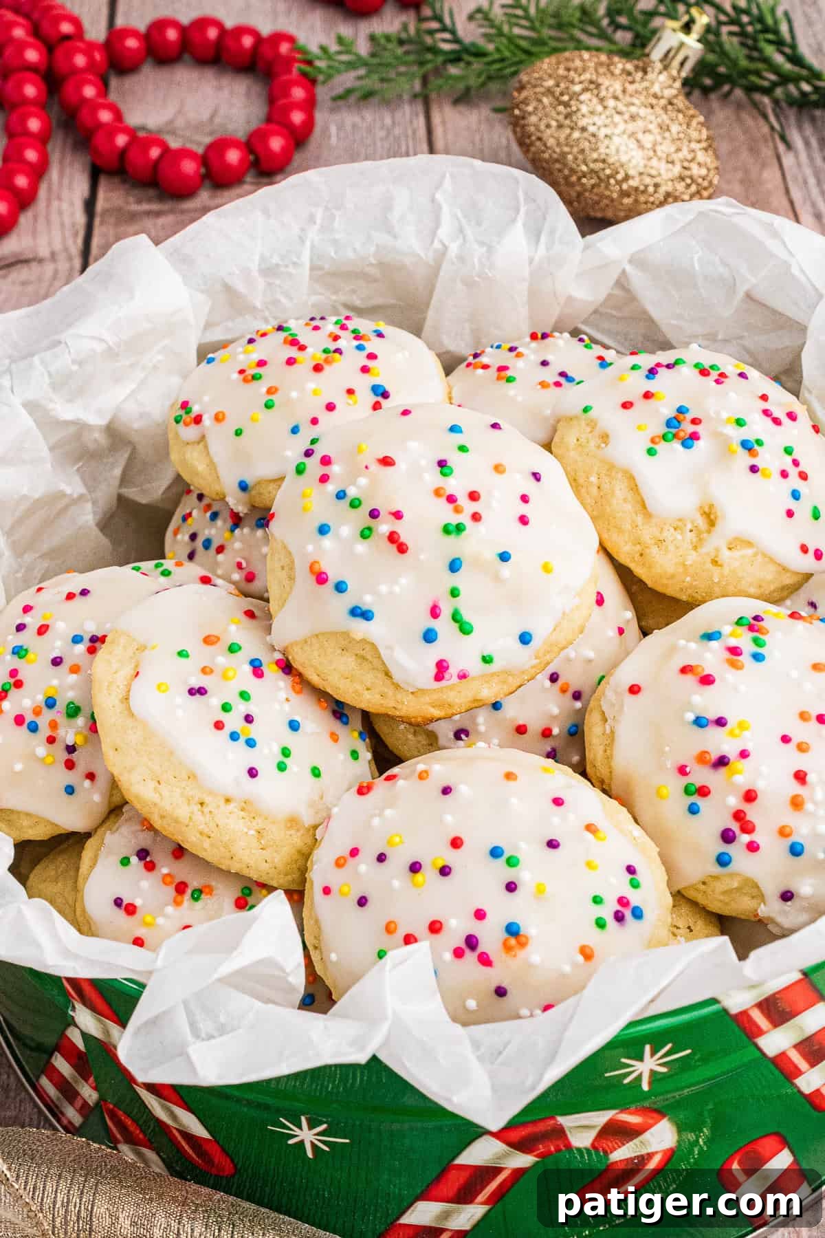 Classic Italian Ricotta Cookies 2 A festive display of Italian ricotta cookies with white icing and colorful sprinkles, piled in a holiday-themed tin lined with parchment paper. The background features red wooden beads, a gold ornament, and green pine branches, enhancing the Christmas setting.