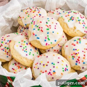 Classic Italian Ricotta Cookies 10 A close-up of Italian ricotta cookies topped with smooth white icing and colorful round sprinkles, arranged in a festive green tin lined with parchment paper. The cookies are soft, fluffy, and stacked to showcase their homemade charm.
