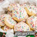 Classic Italian Ricotta Cookies 11 A close-up of Italian ricotta cookies topped with smooth white icing and colorful round sprinkles, arranged in a festive green tin lined with parchment paper. The cookies are soft, fluffy, and stacked to showcase their homemade charm.
