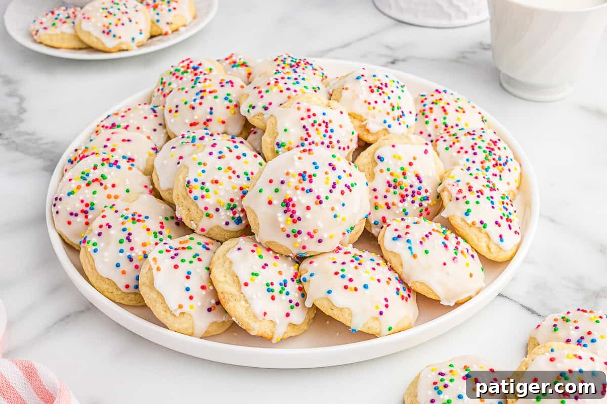 Classic Italian Ricotta Cookies 3 A white plate piled high with Italian ricotta cookies topped with smooth white icing and colorful sprinkles. Additional cookies are visible on a smaller plate in the background, set on a marble surface, creating a bright and festive display.