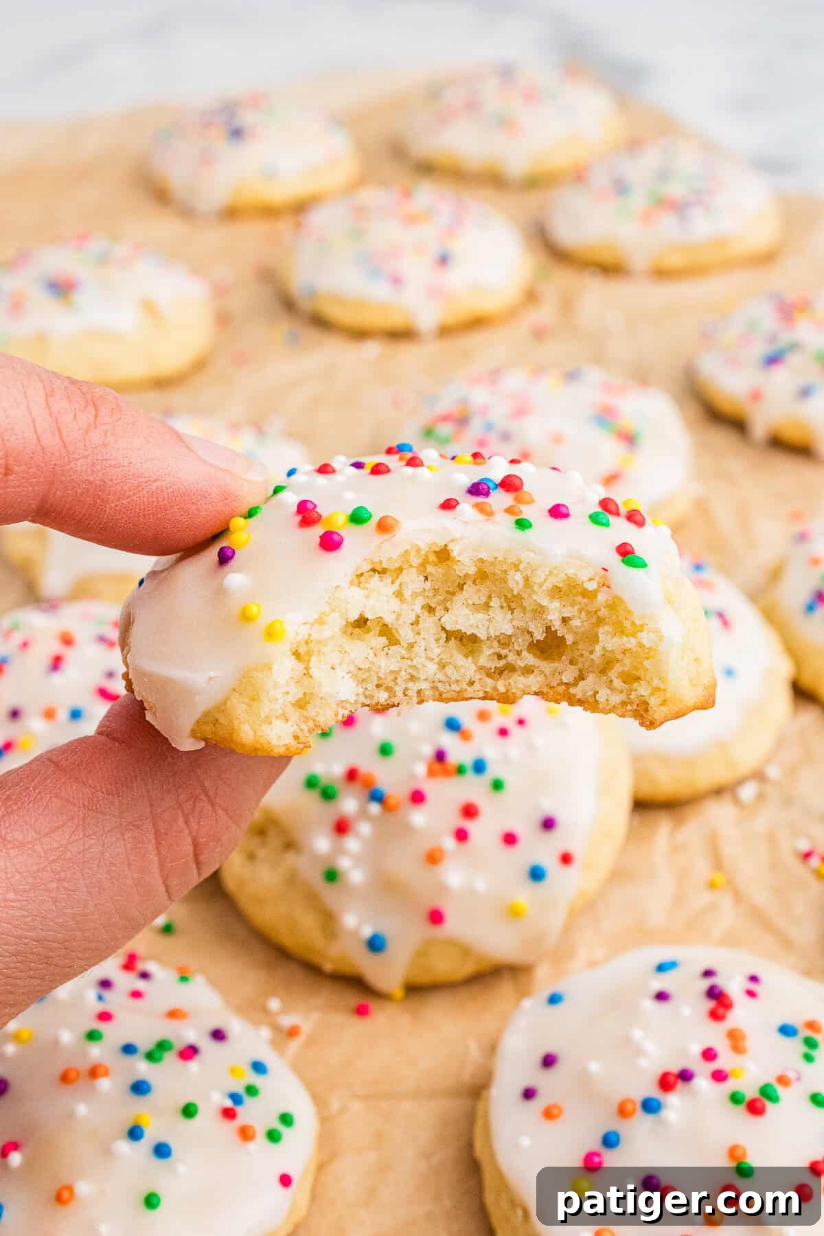 Classic Italian Ricotta Cookies 9 A hand holds a half-eaten Italian ricotta cookie with white icing and colorful sprinkles, revealing its soft, fluffy interior. More cookies are scattered in the background on parchment paper, highlighting the festive and homemade presentation.