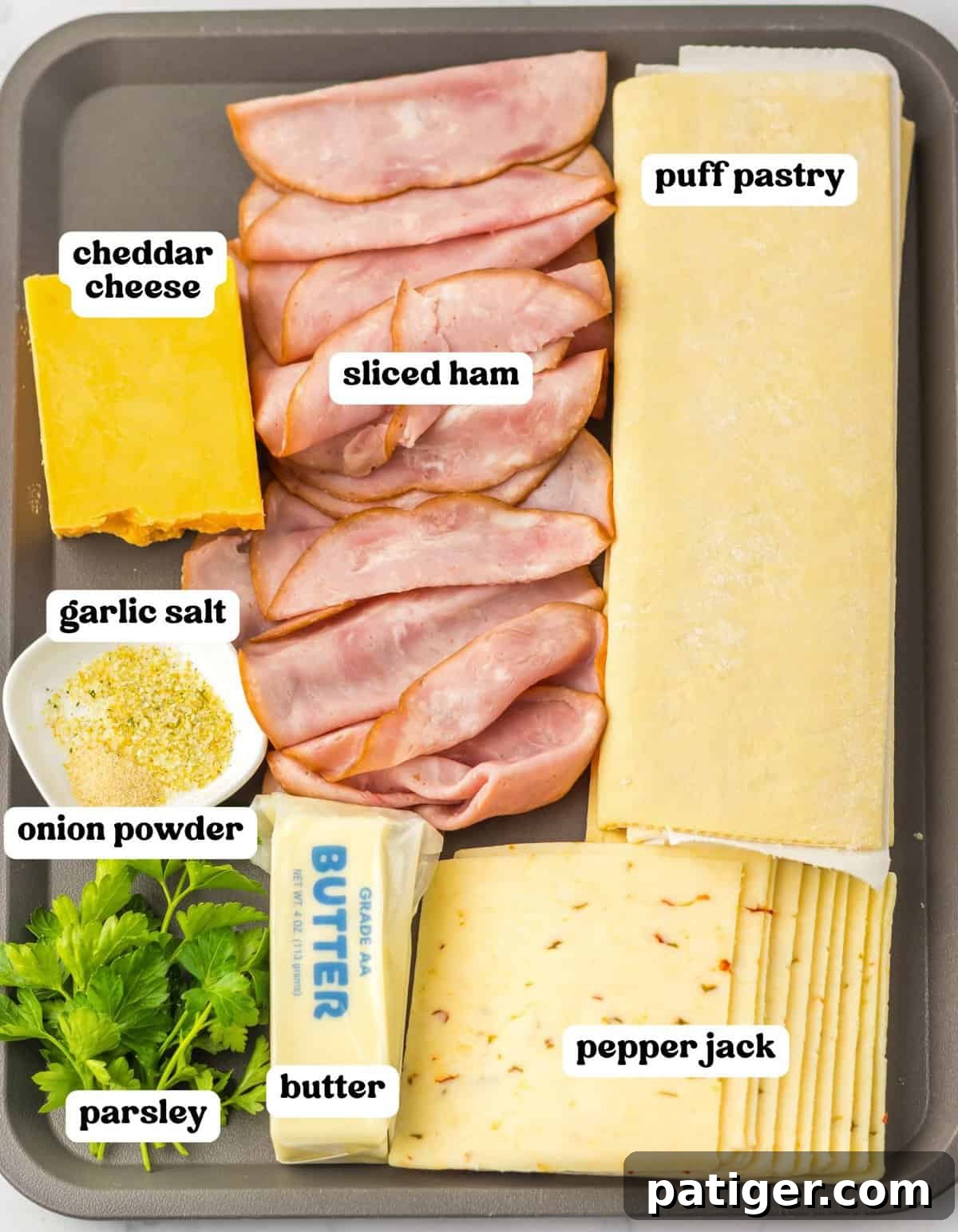 Tray with ingredients for puff pastry Christmas trees: sliced ham, cheddar cheese, pepper jack cheese, puff pastry sheets, butter, parsley, garlic salt, and onion powder.